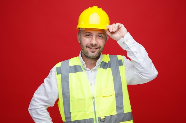 Foto hombre constructor en uniforme de construcción y casco de seguridad mirando a la cámara feliz y positivo saludo sonriente de pie sobre fondo rojo.