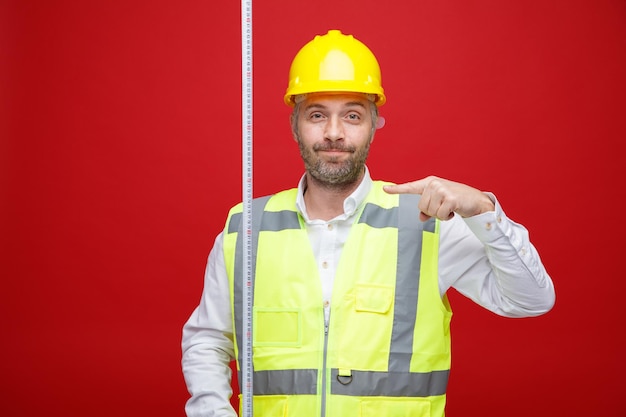 Hombre constructor en uniforme de construcción y casco de seguridad sosteniendo cinta métrica apuntando con el dedo índice sonriendo mirando a la cámara de pie sobre fondo rojo.