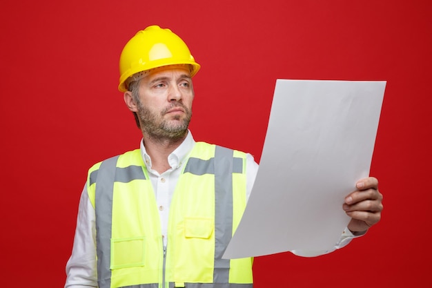 Hombre constructor en uniforme de construcción y casco de seguridad sosteniendo un plan mirándolo con cara seria de pie sobre fondo rojo.