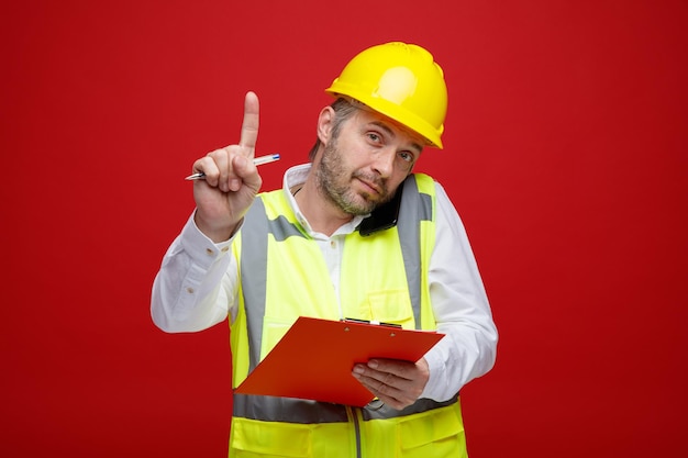 Hombre constructor en uniforme de construcción y casco de seguridad sosteniendo portapapeles hablando por teléfono móvil mostrando el dedo índice como pedir un minuto de pie sobre fondo rojo