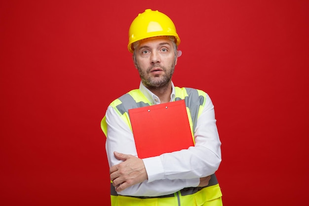 Hombre constructor en uniforme de construcción y casco de seguridad sosteniendo portapapeles mirando a la cámara con cara seria de pie sobre fondo rojo.