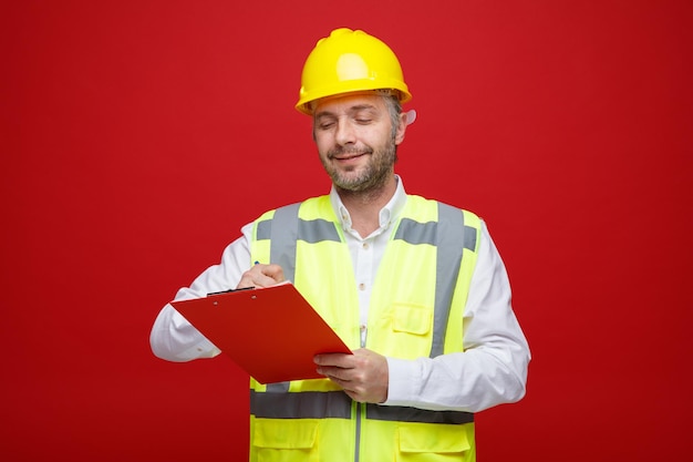 Hombre constructor en uniforme de construcción y casco de seguridad sosteniendo portapapeles mirando confiado sonriendo haciendo notas de pie sobre fondo rojo.