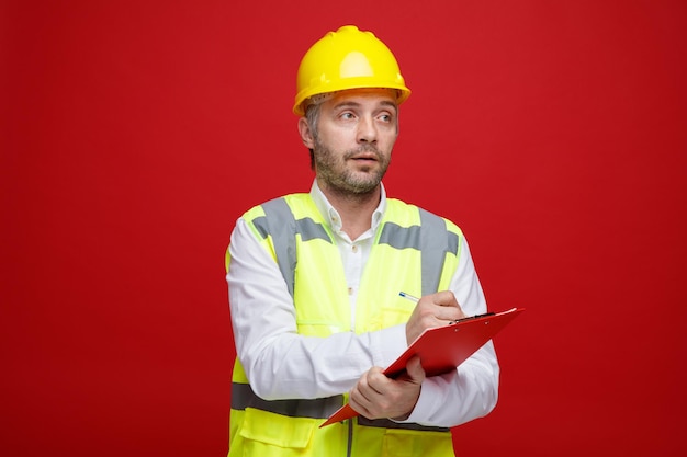 Hombre constructor en uniforme de construcción y casco de seguridad sosteniendo portapapeles mirando a un lado con cara seria haciendo notas de pie sobre fondo rojo.