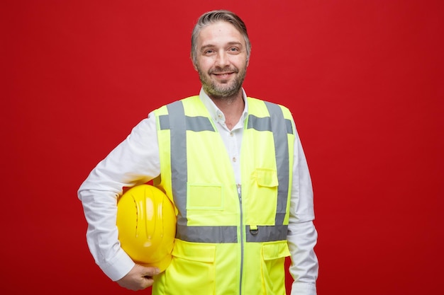 Hombre constructor en uniforme de construcción sosteniendo su casco de seguridad mirando a la cámara feliz y confiado sonriendo de pie sobre fondo rojo.