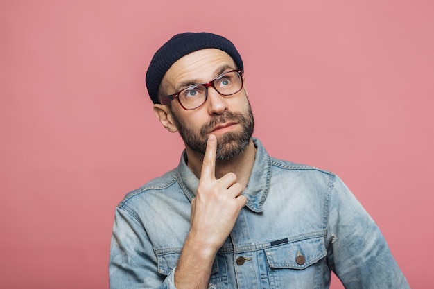 Hombre contemplativo con barba tocando su barbilla en pensamiento con gafas rojas y una chaqueta de denim contra un fondo rosa vibrante