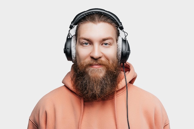 Un hombre contento y apuesto escandinavo busca sonrisas y escucha música en auriculares profesionales aislados en un fondo gris Chico feliz con barba y peinado de jengibre Tecnologías digitales modernas