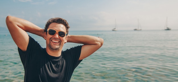 Un hombre contento con gafas de sol con las manos detrás de la cabeza junto al agua.