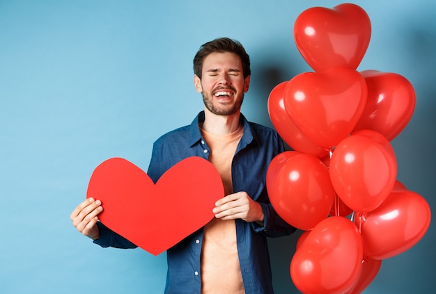 Foto hombre con el corazón roto llorando por la ruptura del día de san valentín, sosteniendo un recorte de corazón rojo y parado cerca de globos románticos sobre fondo azul.