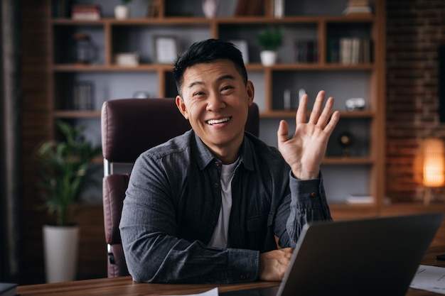 Hombre coreano adulto feliz en el lugar de trabajo con la computadora portátil mira la cámara agitando la mano en el interior de la oficina en casa