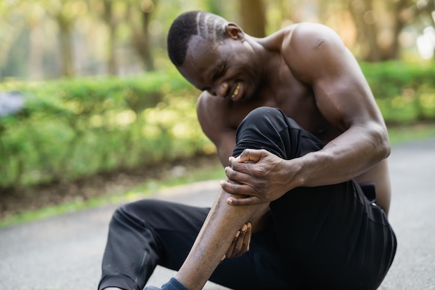Hombre corredor africano sentado en la pista de jogging y sintiendo dolor en su espacio de copia de la pierna