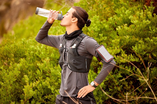 Hombre corredor fitness y agua potable al aire libre para caminatas, entrenamiento cardiovascular o ejercicio. Descanso deportivo de atletas con bebida de botella y bienestar deportivo corriendo en un parque natural o senderismo de montaña.