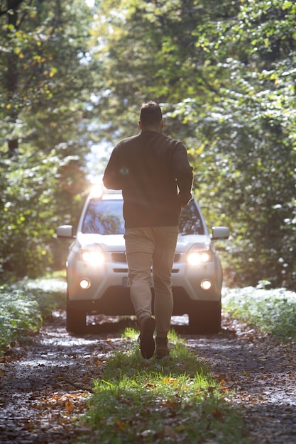 Hombre corriendo al coche parado en el sendero del bosque con los faros encendidos