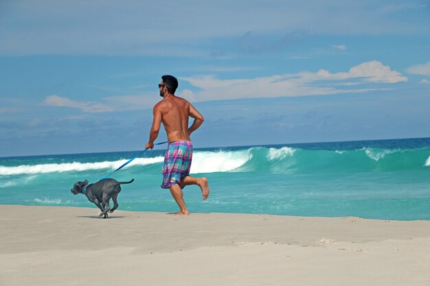 Hombre corriendo en la playa con su perro pit bull. Día soleado, con cielo azul y mar cristalino.