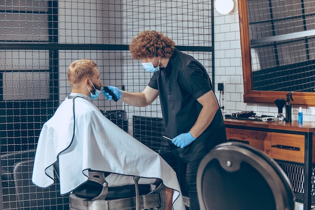 Hombre cortándose el pelo en la barbería con máscara durante la pandemia de coronavirus. Peluquero profesional con guantes. Covid-19, concepto de belleza, cuidado personal, estilo, salud y medicina.