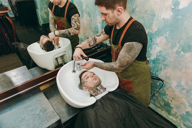 Hombre de corte de pelo de moda en la peluquería. El estilista masculino en tatuajes al servicio del cliente, lavándose la cabeza