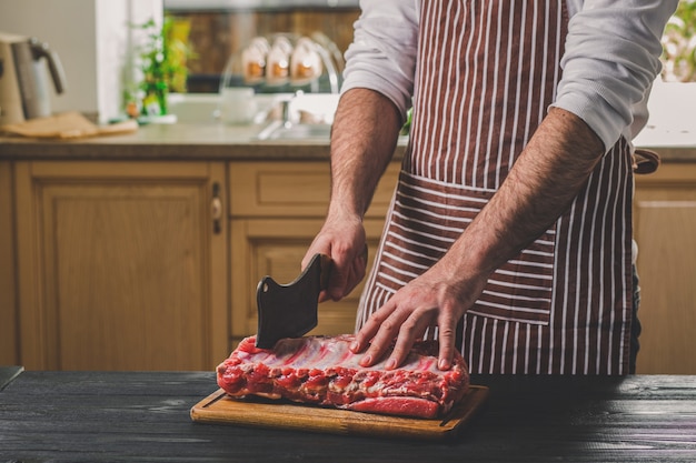 Hombre cortes de trozo de carne fresca sobre una tabla de cortar de madera en la cocina de casa. Un hombre con un delantal a rayas con un gran cuchillo en sus manos