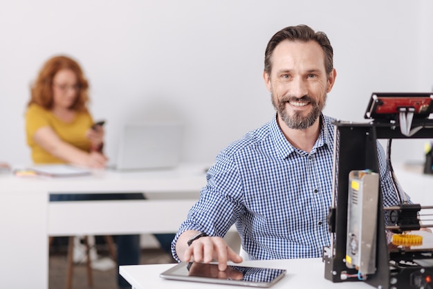 Hombre creativo alegre feliz usando una tableta y buscando nuevas ideas para su proyecto mientras te mira