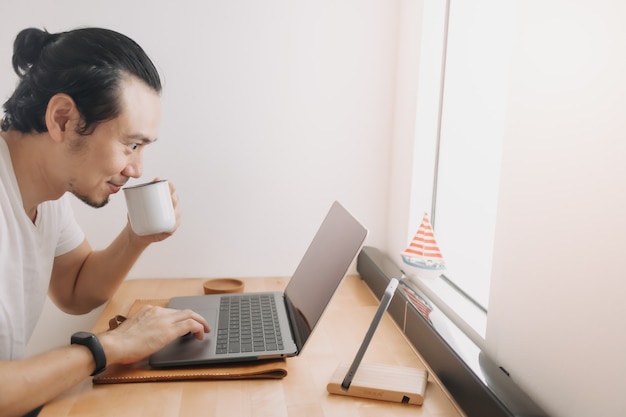 Hombre creativo trabaja con ordenador portátil en el espacio de trabajo de escritorio de madera junto a las ventanas