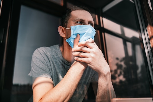 El hombre en cuarentena debido a un virus se sienta en su casa con una máscara y mira por la ventana en un día soleado. Concepto de coronavirus. COVID-19