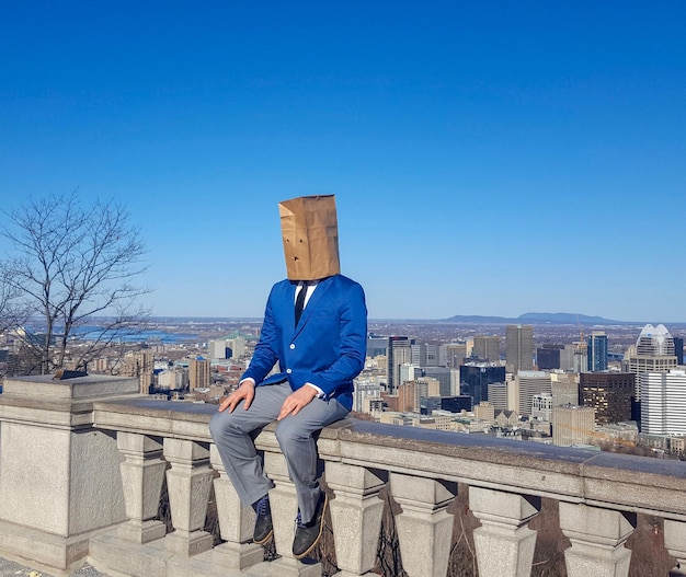 Hombre cubierto con una bolsa de papel sentado en la barandilla contra el cielo azul claro