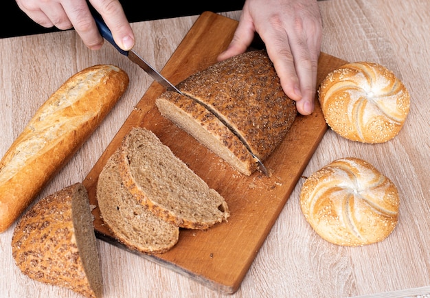 Un hombre con un cuchillo corta el pan sobre una tabla de madera. Baguettes francesas. Diferentes razas sobre fondo de madera.