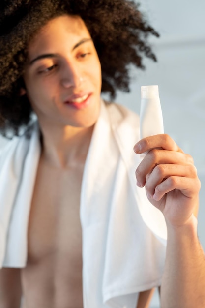 Hombre cuidado de la piel Publicidad de productos cosméticos Tratamiento facial Alegre guapo sin camisa marrón toalla de pelo rizado después de la ducha sosteniendo un tubo de crema de maqueta blanca en la mano sobre un fondo claro desenfocado