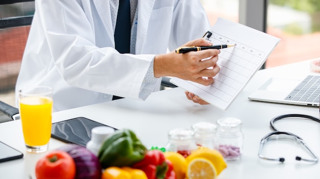 Hombre de cultivo en uniforme médico sentado en el escritorio con alimentos saludables y vitaminas y tomando notas en la mesa durante el trabajo en el hospital