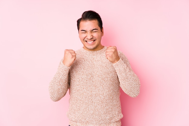 Hombre con curvas joven que presenta en una pared rosada aislada levantando el puño, sintiéndose feliz y acertado. Concepto de victoria