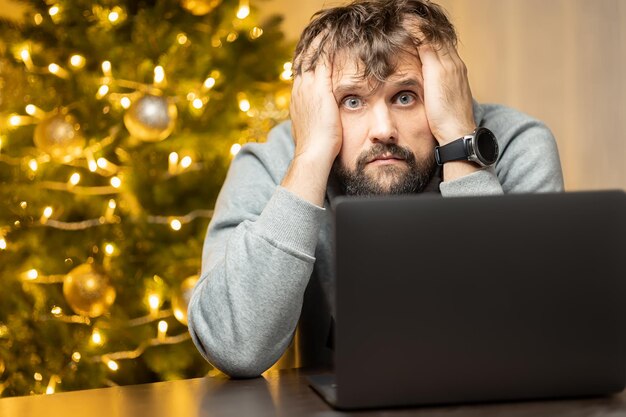 Un hombre cuyo shaggy cansado se sienta en una computadora portátil en la noche de Navidad