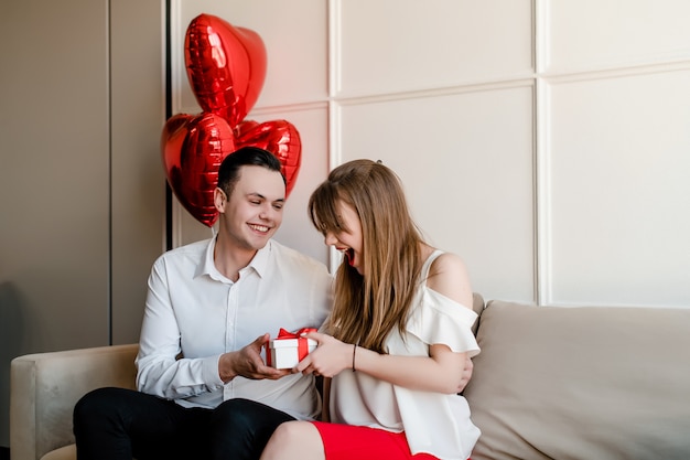 El hombre da un regalo a la mujer sonriente con globos en forma de corazón en el sofá en casa