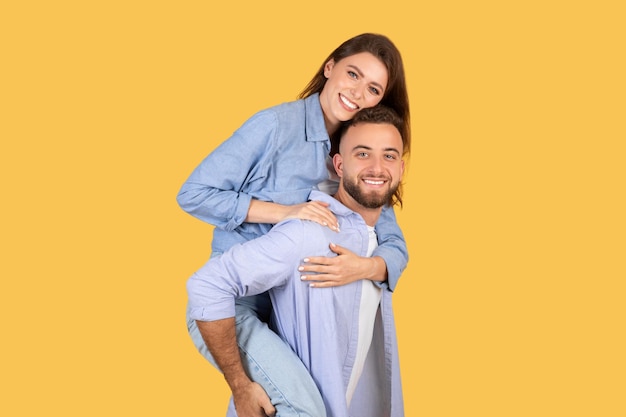 Hombre dando paseo a cuestas a la mujer ambos sonriendo alegremente sobre fondo amarillo