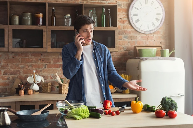 Hombre decepcionado cocinando comida sana y llamando por teléfono en la cocina del desván en casa en un día soleado. Preparando ensalada de verduras.