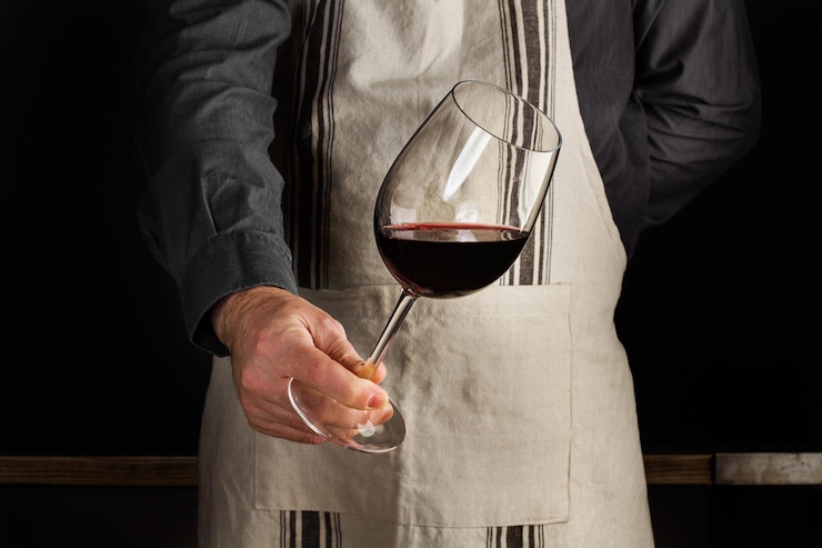 Un hombre con un delantal con una copa de vino en la mano sobre un fondo oscuro