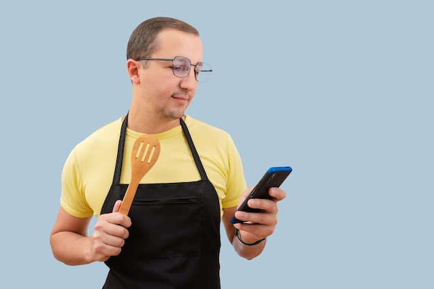 Un hombre en un delantal con una cuchara de madera y un teléfono en sus manos aislado en un fondo azul.