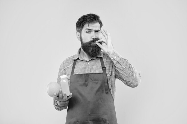 Foto hombre en delantal lamiendo dedo sosteniendo naranja y botella de jugo fondo amarillo jugo barman