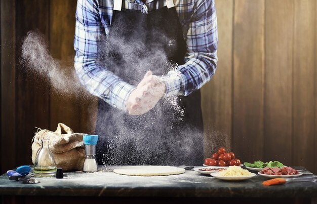 Hombre en un delantal preparando una pizza, amasa la masa y pone los ingredientes