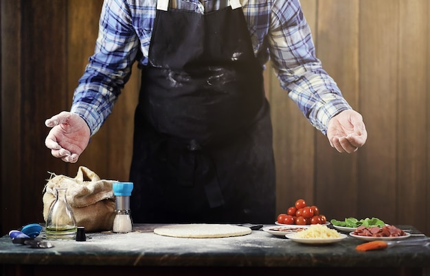 Hombre en un delantal preparando una pizza, amasa la masa y pone los ingredientes