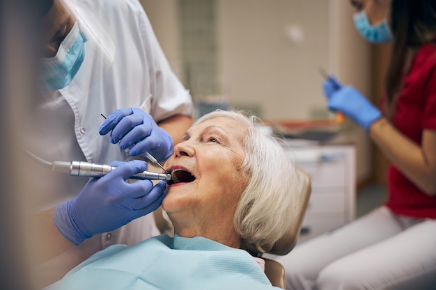 Hombre dentista con herramientas dentales y taladro mientras cuida los dientes del paciente en la oficina de la clínica dental