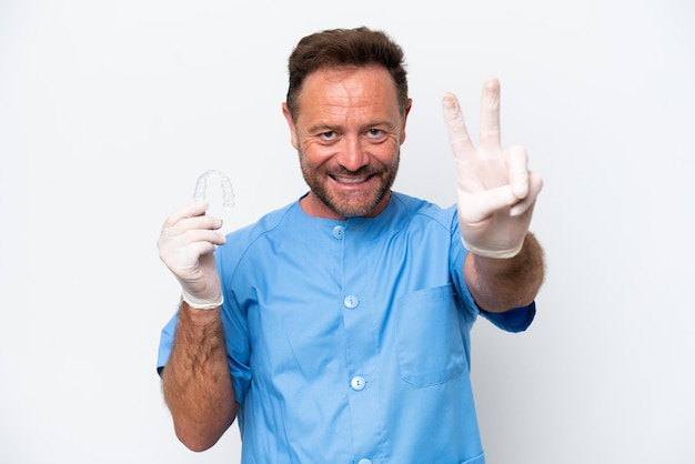 Hombre dentista de mediana edad sosteniendo previendo aislado sobre fondo blanco sonriendo y mostrando el signo de la victoria