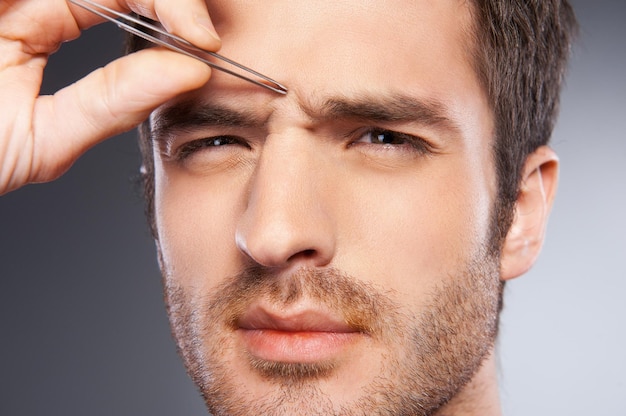 Foto hombre depilarse las cejas. hombre joven frustrado depilarse las cejas y mirando a la cámara mientras está de pie aislado sobre fondo gris