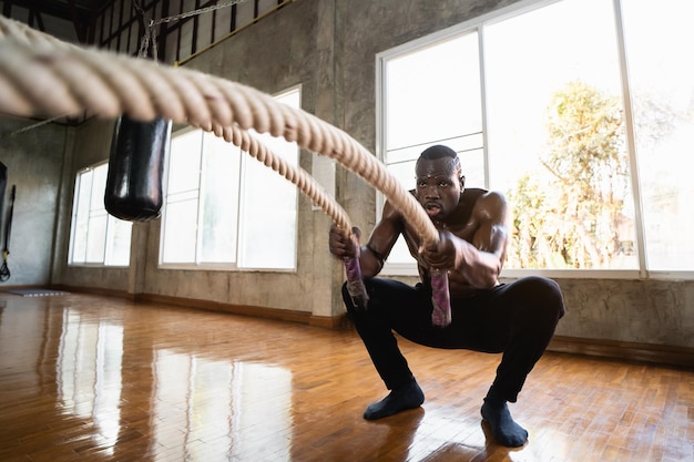 Hombre de deporte africano haciendo entrenamiento funcional de cuerdas de batalla en el gimnasio. Entrenador decidido haciendo olas con cuerdas mientras ejercita fuerza. Atleta trabajando con cuerda de batalla en el gimnasio industrial.