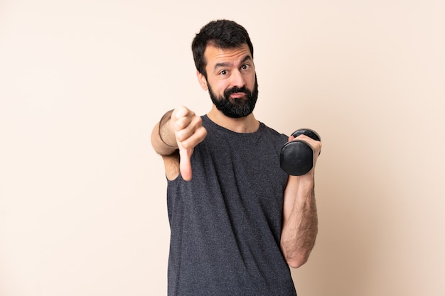 Hombre de deporte caucásico con barba haciendo levantamiento de pesas sobre fondo aislado mostrando el pulgar hacia abajo con expresión negativa