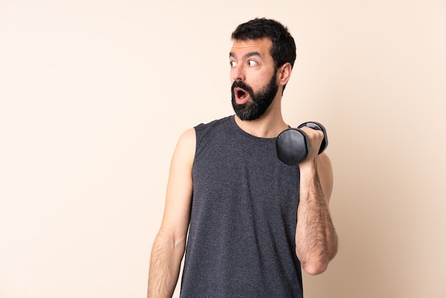 Hombre de deporte caucásico con barba haciendo levantamiento de pesas sobre pared aislada haciendo gesto de sorpresa mientras mira hacia el lado