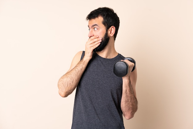 Hombre de deporte caucásico con barba haciendo levantamiento de pesas sobre pared aislada que cubre la boca y mirando hacia el lado