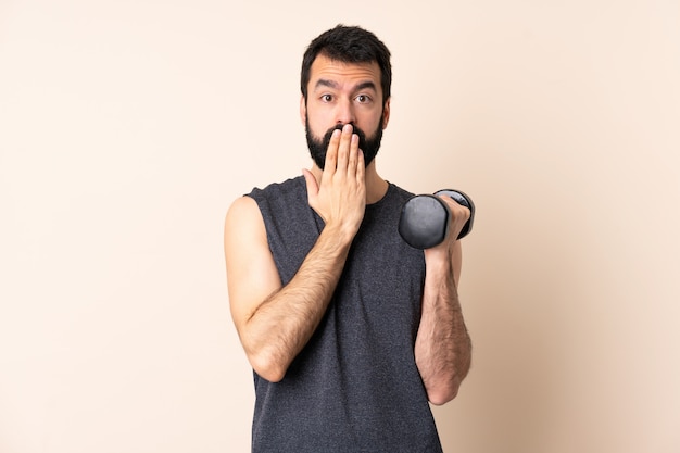 Hombre de deporte caucásico con barba haciendo levantamiento de pesas sobre la pared que cubre la boca con la mano