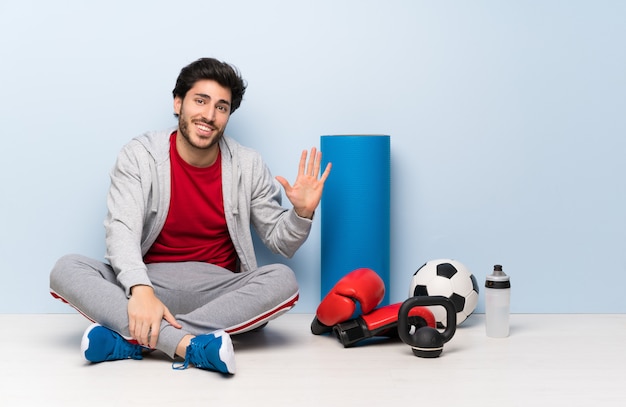 Hombre del deporte que se sienta en el piso que saluda con la mano con la expresión feliz