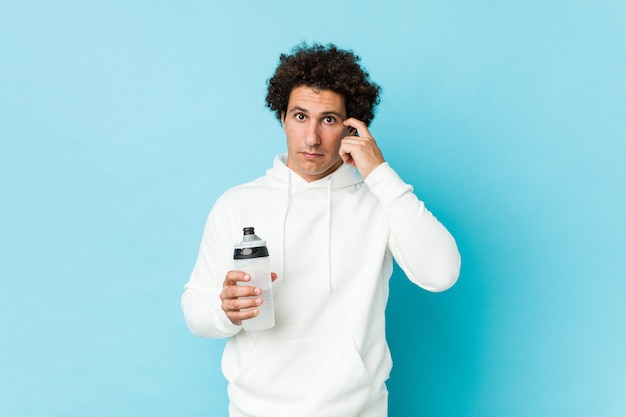 Hombre del deporte que sostiene una botella de agua que señala su sien con el dedo, pensando, centrado en una tarea.