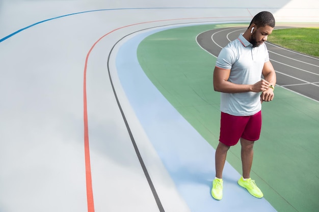 Hombre deportivo afroamericano revisando su rastreador fitbit en la muñeca mientras está de pie en la pista de atletismo