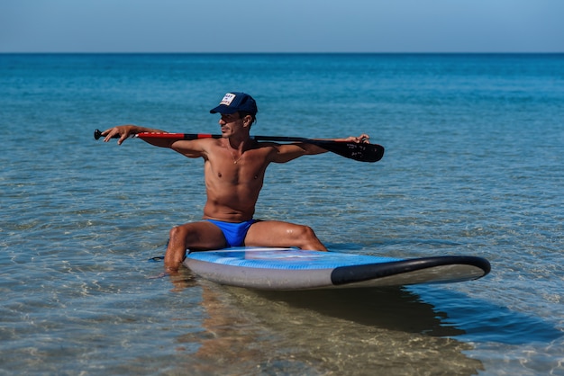 Hombre deportivo bronceado con una gorra se sienta en su tabla de surf en el agua sosteniendo con las manos un remo detrás de la cabeza y mira hacia el agua.