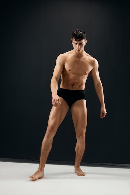 Hombre deportivo de cuerpo completo con cuerpo musculoso en bragas negras posando contra un fondo oscuro Foto de alta calidad